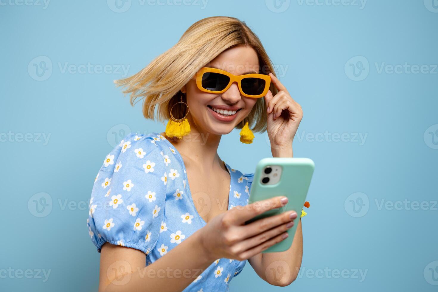 Beautiful young woman using smart phone and smiling against blue background photo