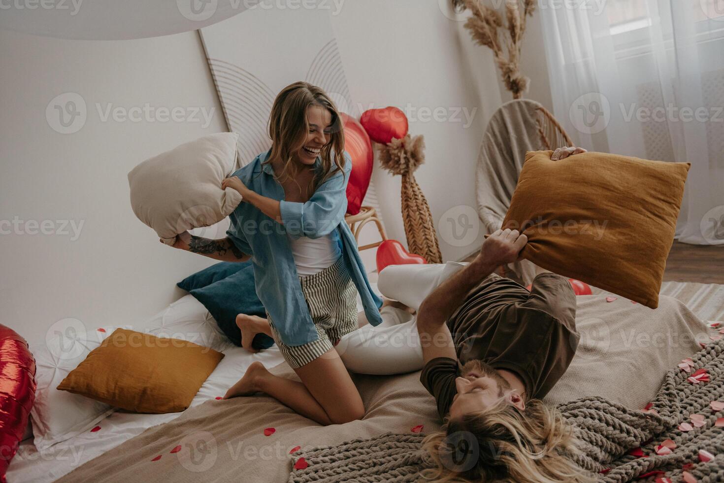 Joyful young loving couple having a pillow fight in bed surrounded with red heart shape balloons photo