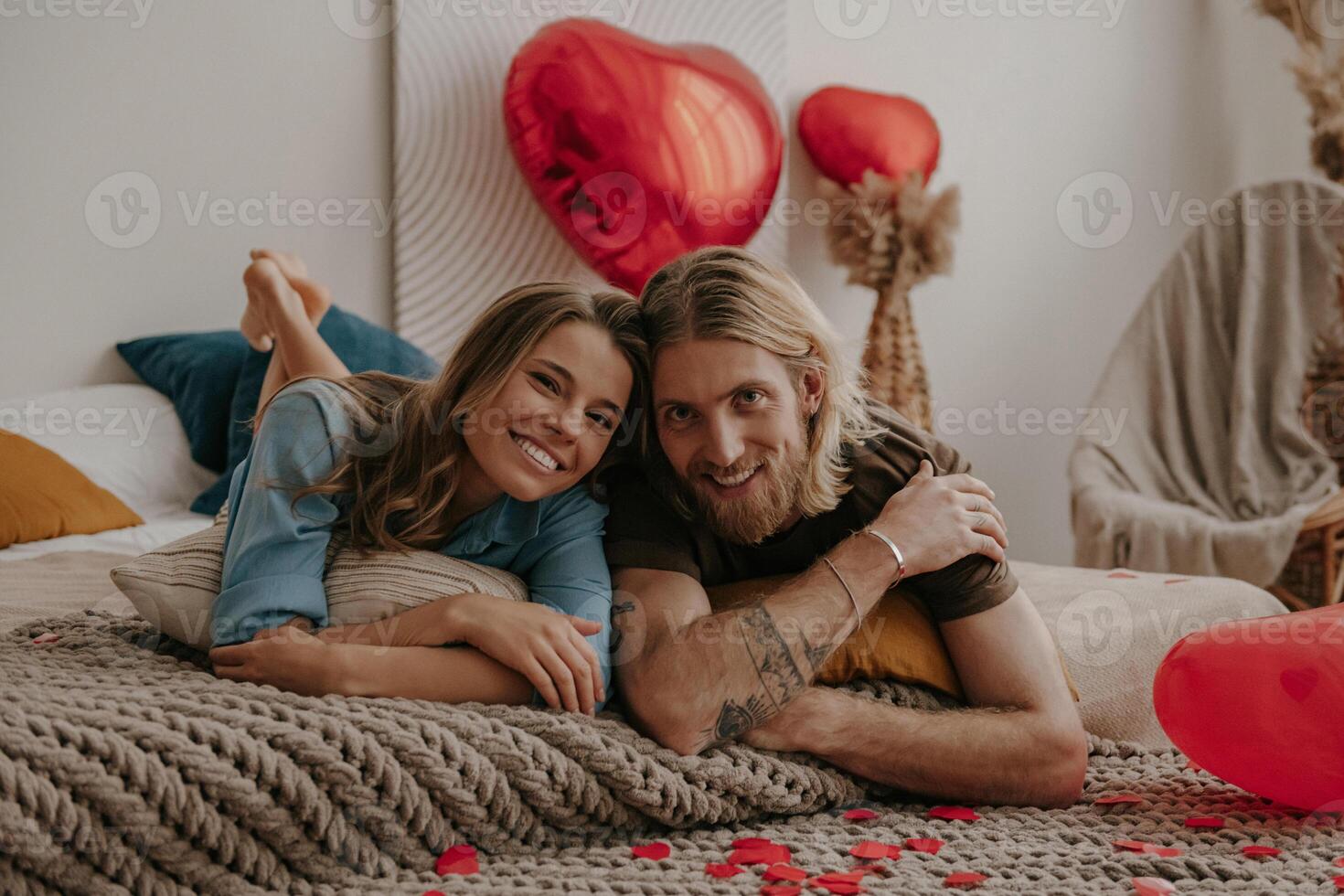Smiling young loving couple lying in bed surrounded with red heart shape balloons and confetti at home photo