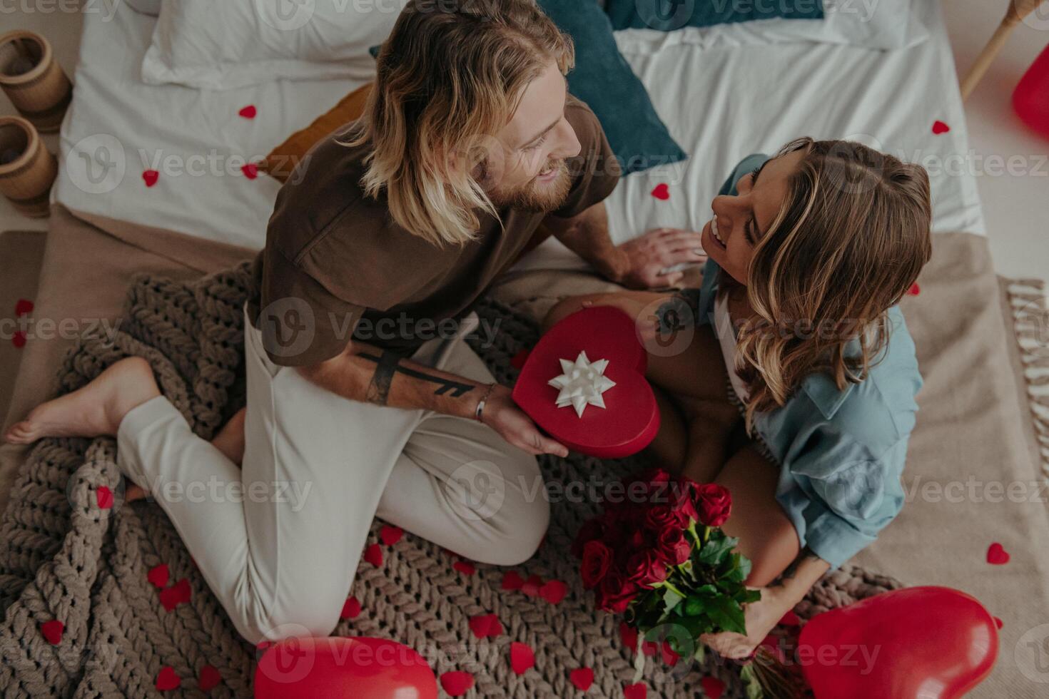 Top view of romantic young man giving a red heart shape gift box to his girlfriend while sitting on bed together photo