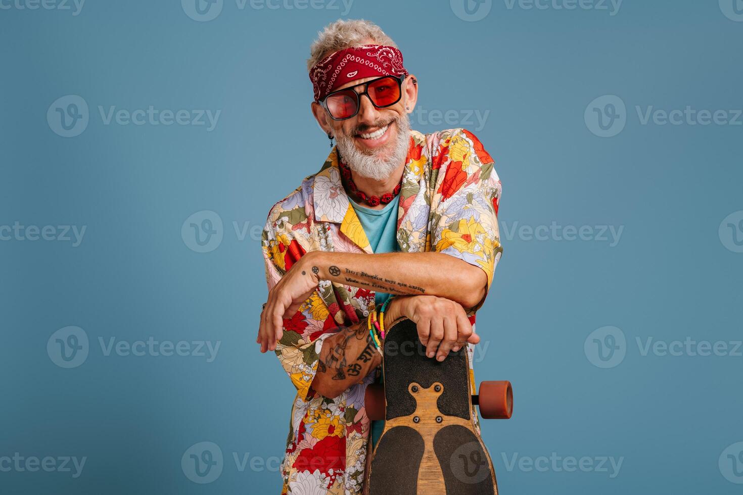 Hipster mature man in stylish funky shirt leaning on longboard and smiling against blue background photo