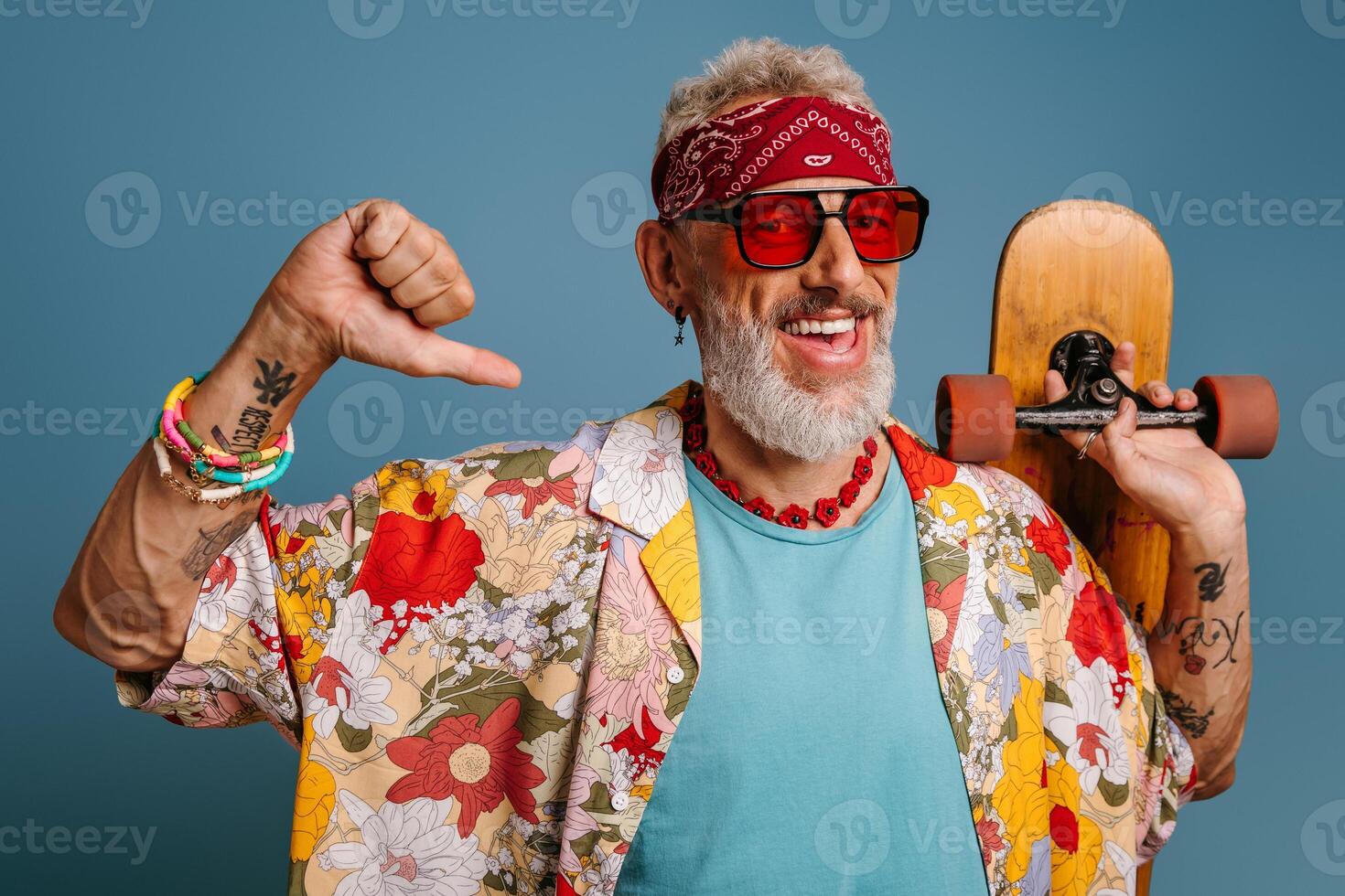 Joyful hipster mature man in stylish funky shirt carrying longboard and pointing self against blue background photo