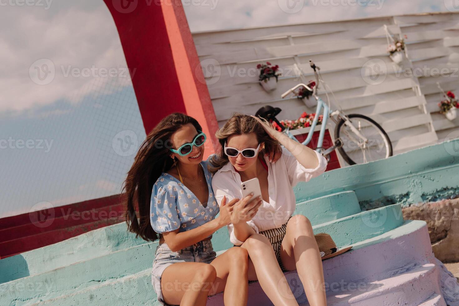 Two attractive young women using smart phone and smiling while sitting outdoors together photo