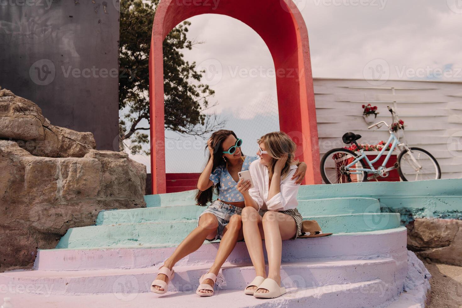 Two happy young women using smart phone while sitting outdoors together photo