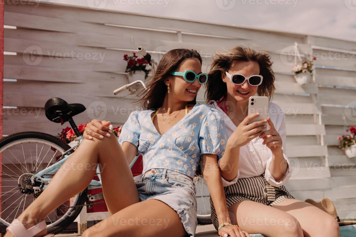 Two attractive young women using smart phone and smiling while sitting outdoors together photo