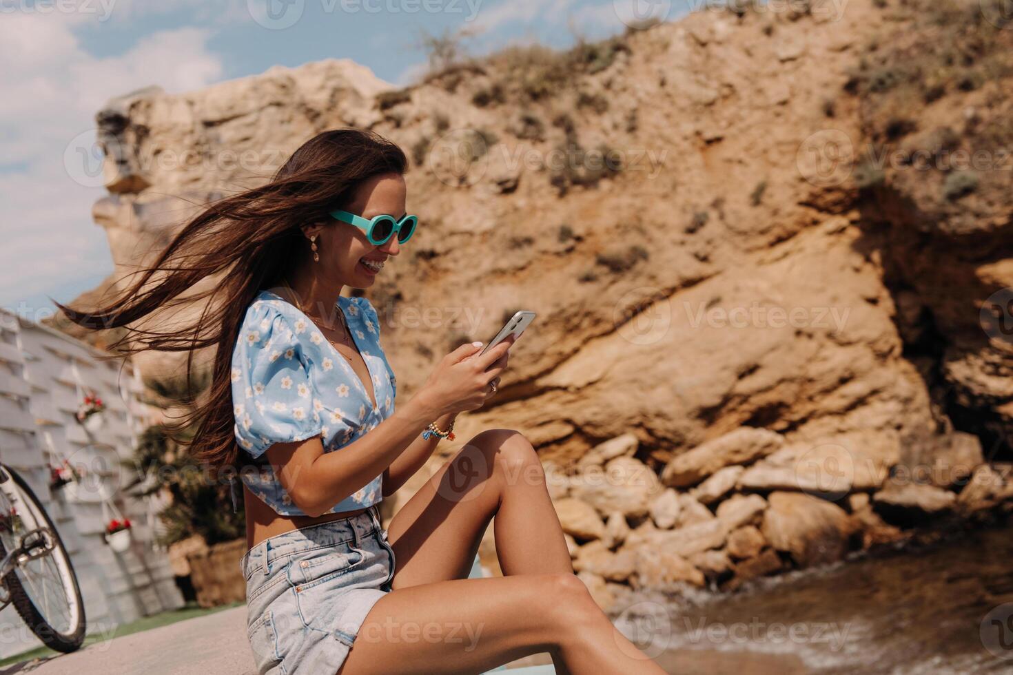 Attractive young woman using smart phone and smiling while sitting seaside photo