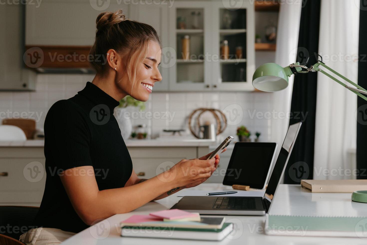 Smiling young woman sitting at the desk and using smart phone while working at home photo