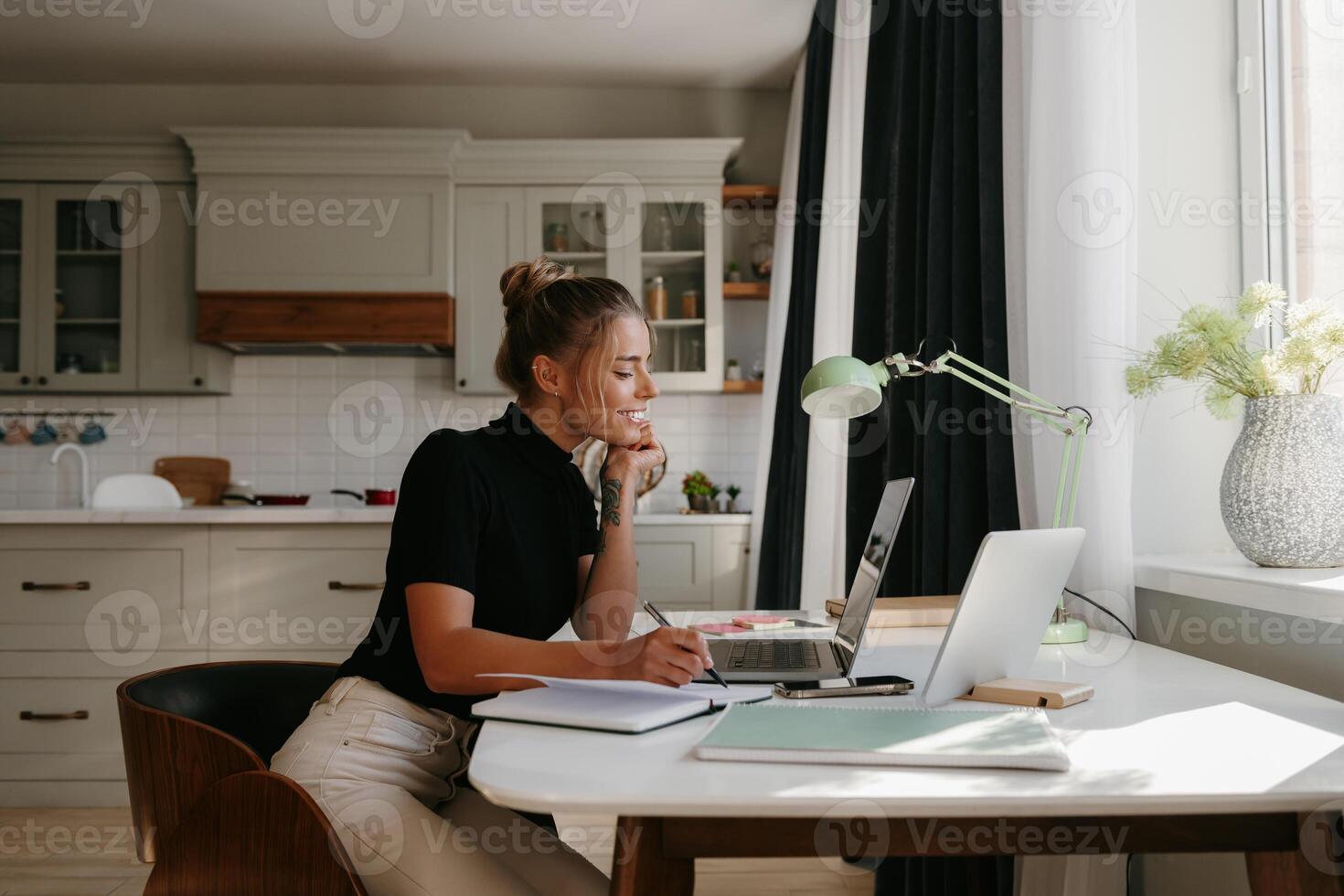 Smiling young woman making notes and looking at the laptop while working at home photo