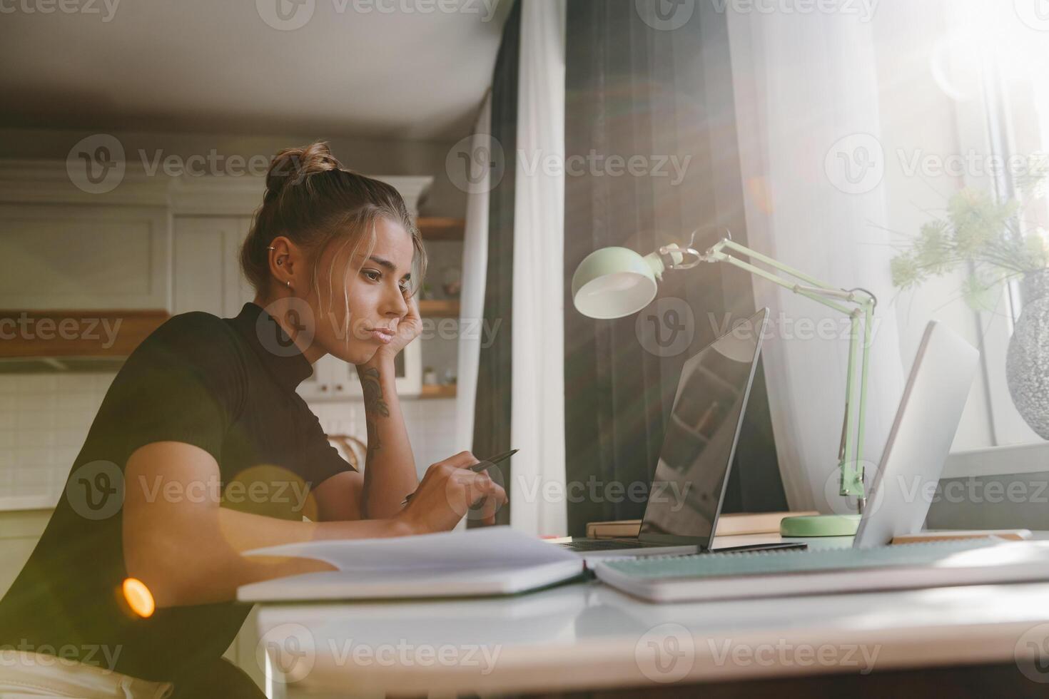 Bored young woman sitting at the desk and looking at laptop while studying at home photo