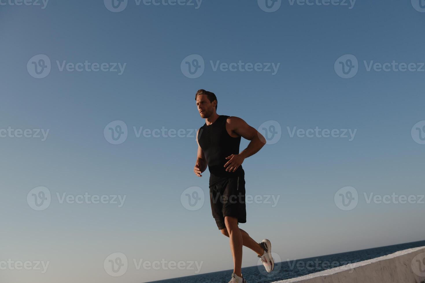 Handsome young man in sports clothing running by the quay with the sea on background photo