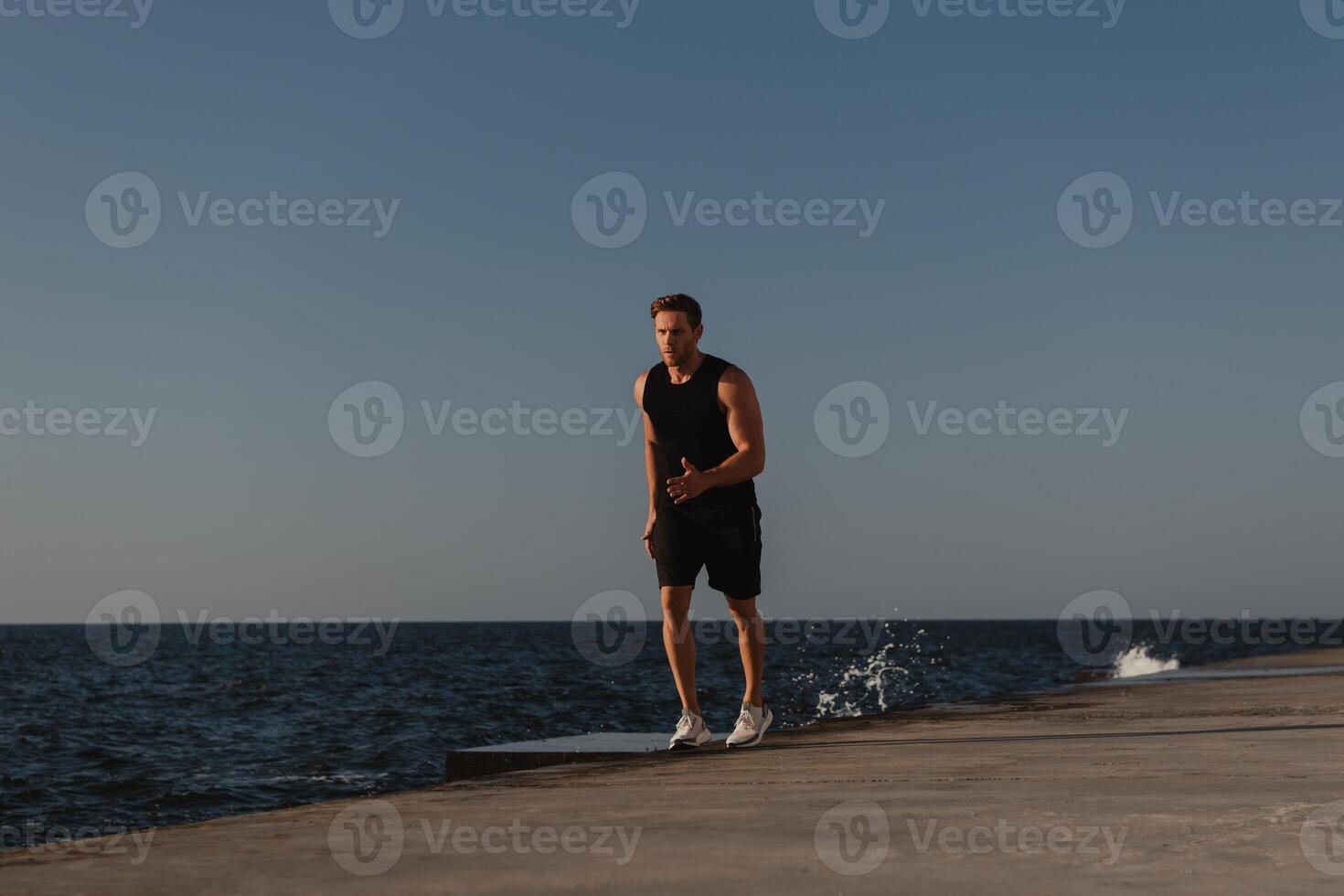 Confident athletic man in sports clothing running by the quay with the sea on background photo