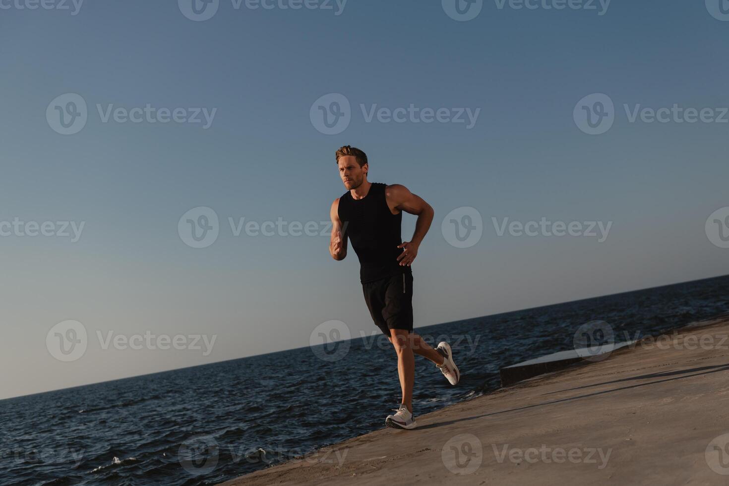 Confident male athlete in sports clothing running by the quay with the sea on background photo