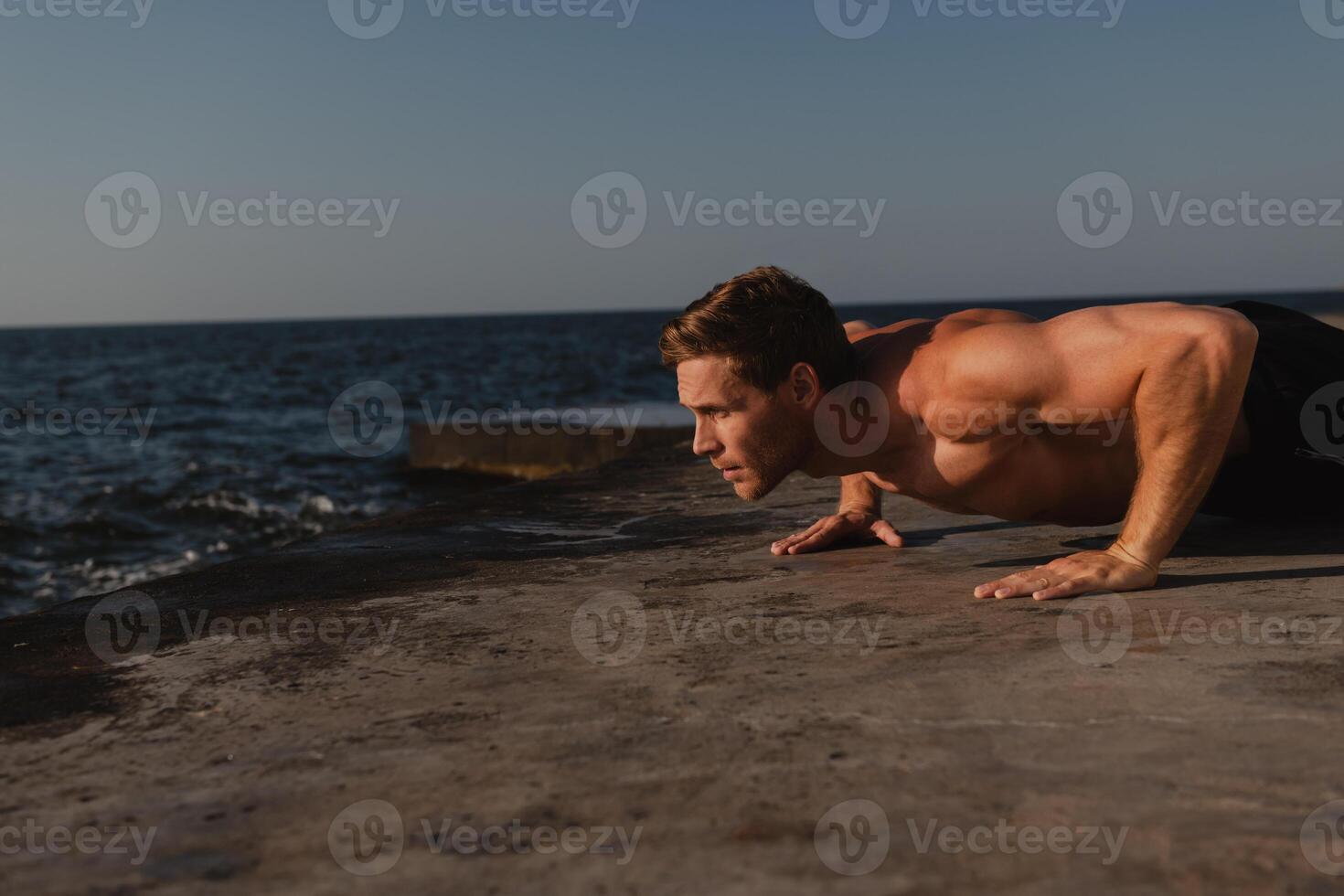 Confident male athlete doing push-ups while exercising outdoors with the sea on background photo