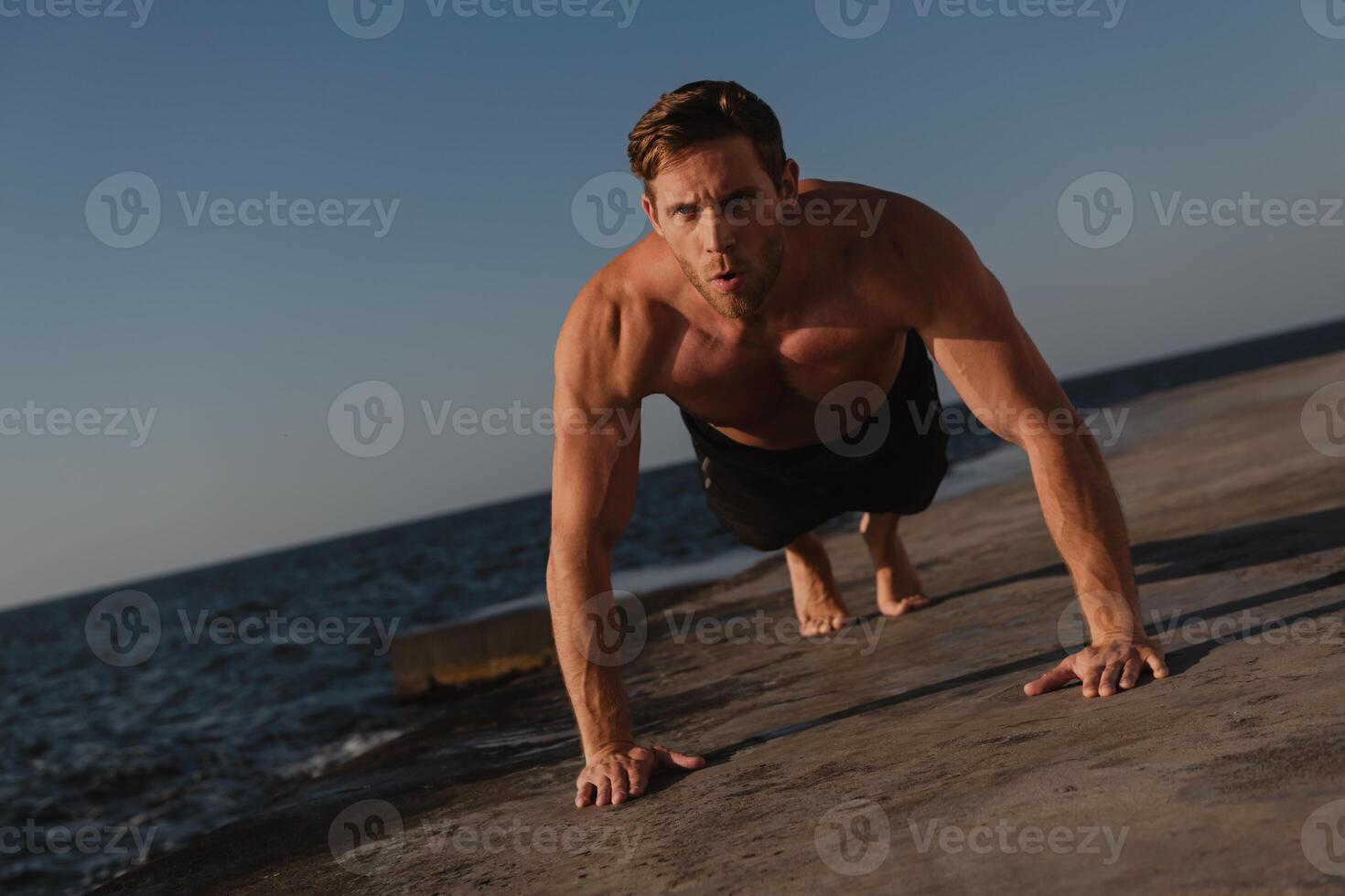 Confident young shirtless man doing push-ups while exercising outdoors with the sea on background photo