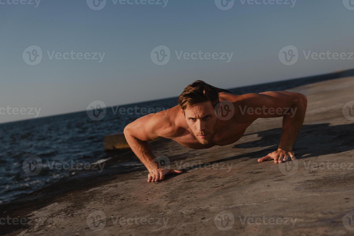 Handsome male athlete doing push-ups while exercising outdoors with the sea on background photo