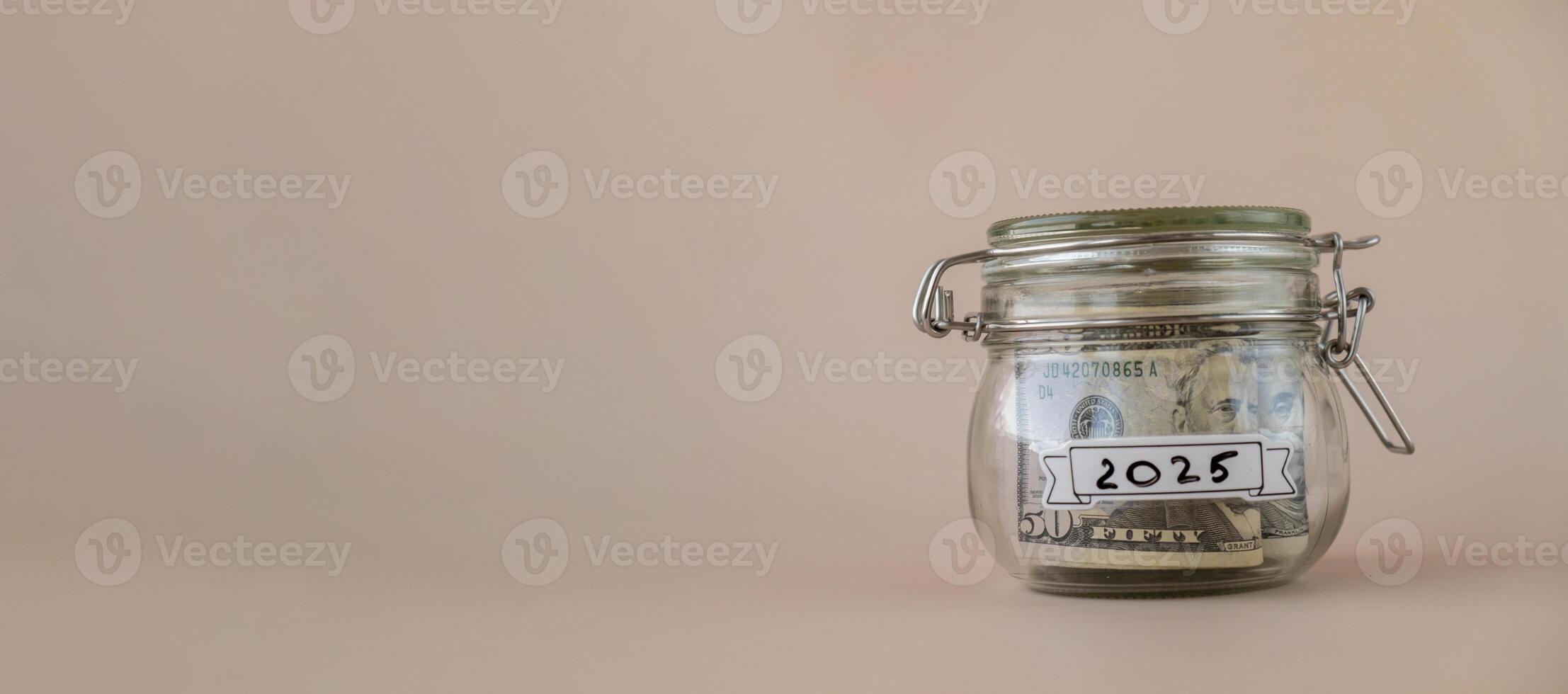Saving Money In Glass Jar filled with Dollars banknotes. 2025 year transcription in front of jar. Managing personal finances extra income for future insecurity. Beige background photo