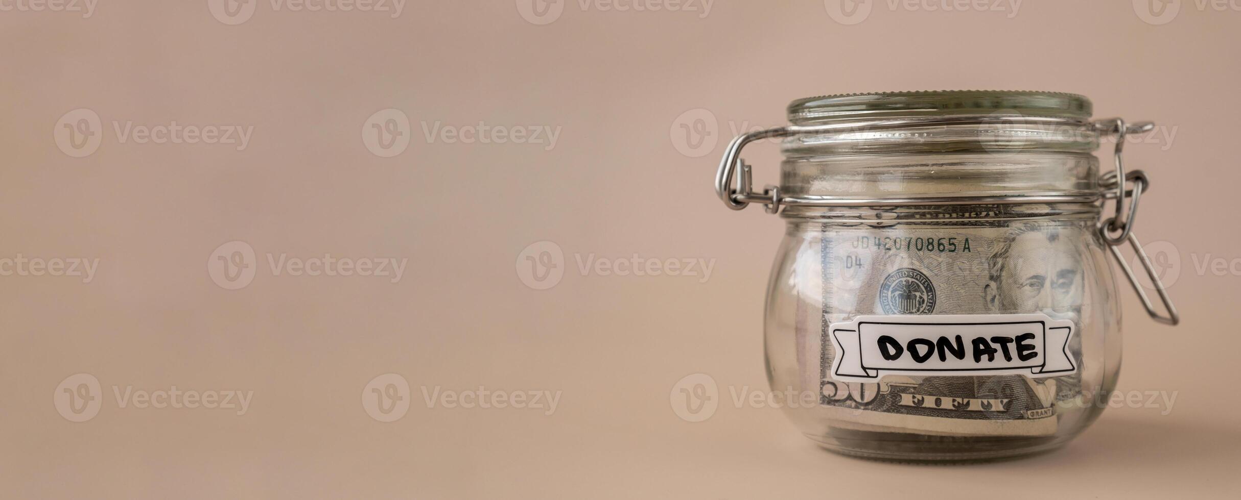 Saving Money In Glass Jar filled with Dollars banknotes. DONATE transcription in front of jar. Managing personal finances extra income for future insecurity. Beige background photo