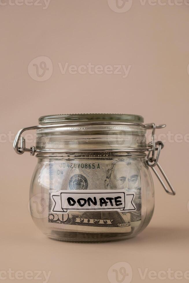 Saving Money In Glass Jar filled with Dollars banknotes. DONATE transcription in front of jar. Managing personal finances extra income for future insecurity. Beige background photo
