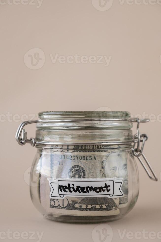 Saving Money In Glass Jar filled with Dollars banknotes. RETIREMENT transcription in front of jar. Managing personal finances extra income for future insecurity. Beige background photo