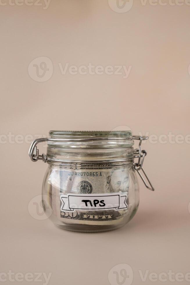 Saving Money In Glass Jar filled with Dollars banknotes. TIPS transcription in front of jar. Managing personal finances extra income for future insecurity. Beige background photo