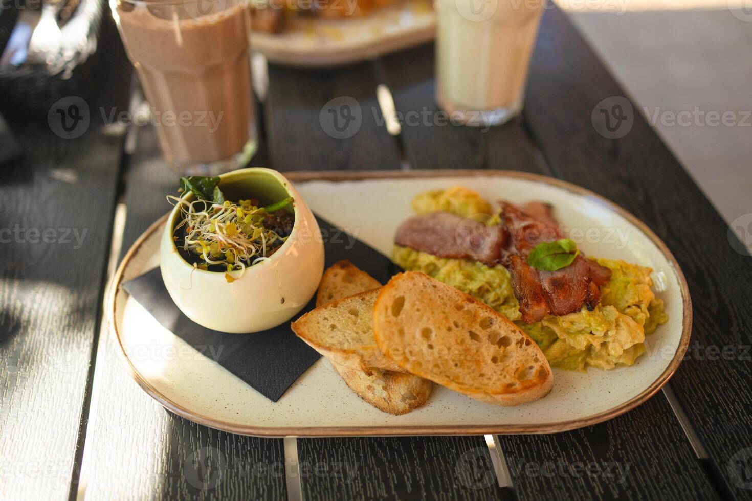 Dish with fried egg, greens, avocado, fried bacon, white crouton bread and pasta. A plate with a classic continental breakfast standing on a wooden table in a restaurant near the window. photo