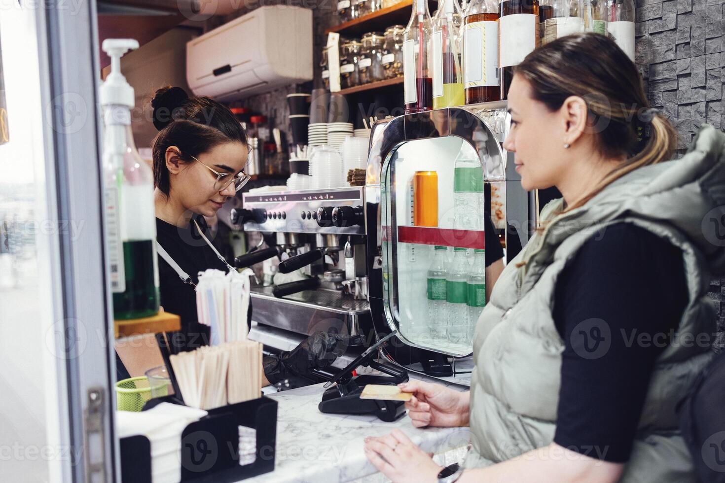 Smiling young barista girl in casual clothes, apron and black gloves accepts order and payment from her customer photo