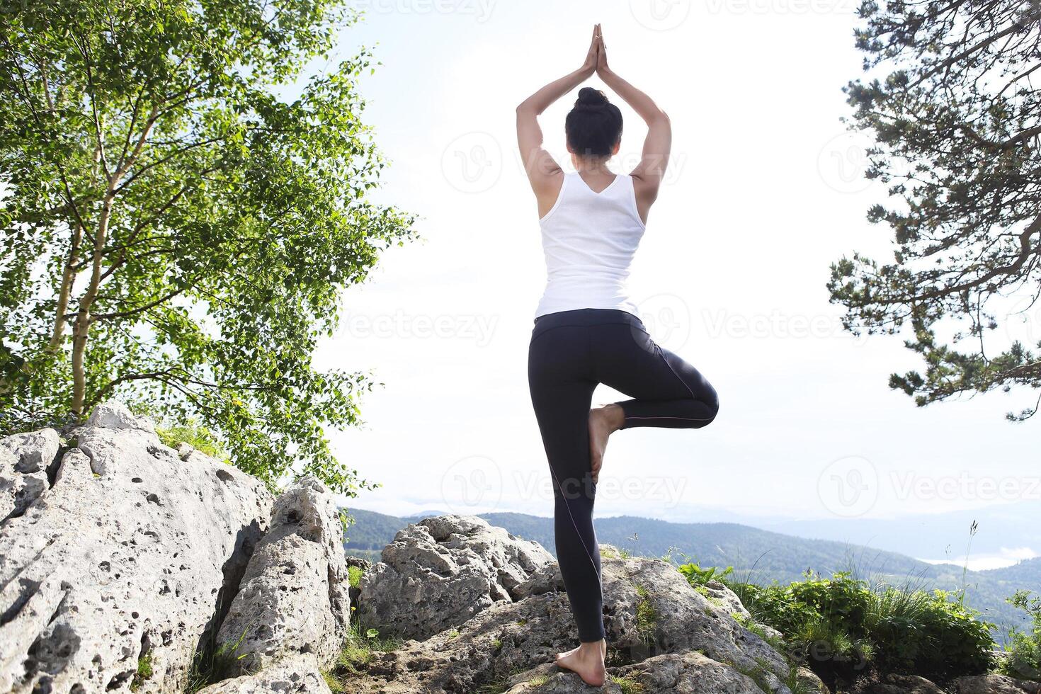 Attractive young woman doing a yoga pose for balance and stretching staying on top of high rock in the mountains photo