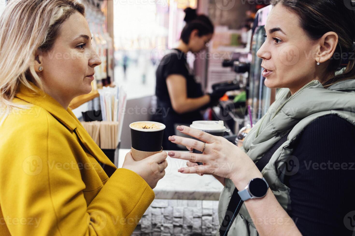 Attractive young women hold paper cups with coffee and chat on front of small street cafe. photo