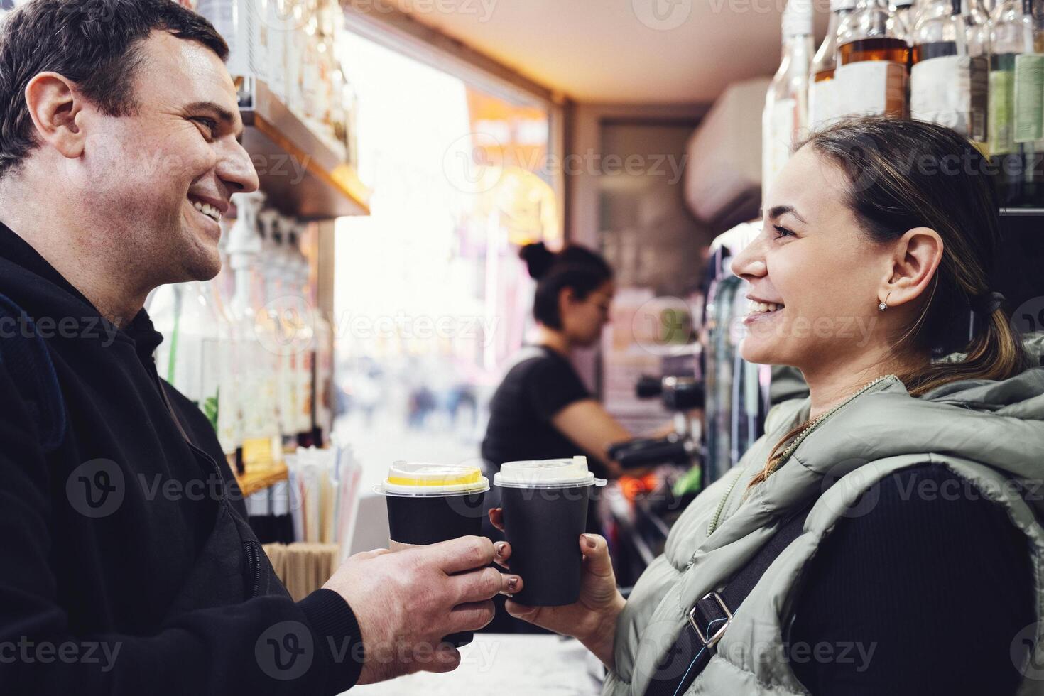 Attractive woman and man holding paper cups with coffee and smiling at each other on front of small street cafe. photo