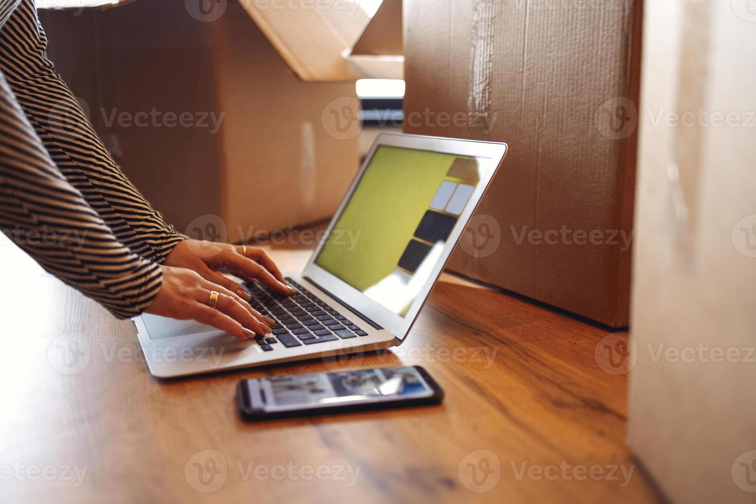 Female hands are typing on a laptop on the parquet floor. There are a telephone nearby and a lot of cardboard boxes with things. The concept of moving from an apartment or house. photo