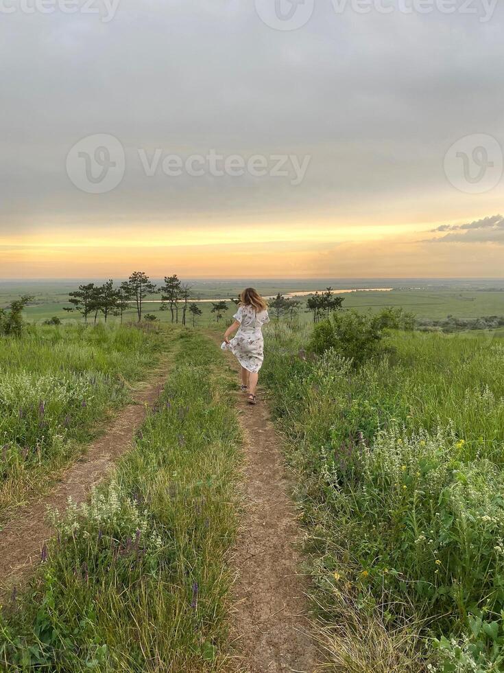 Back view of woman in romantic dress running through field of green grass at sunset on summertime photo