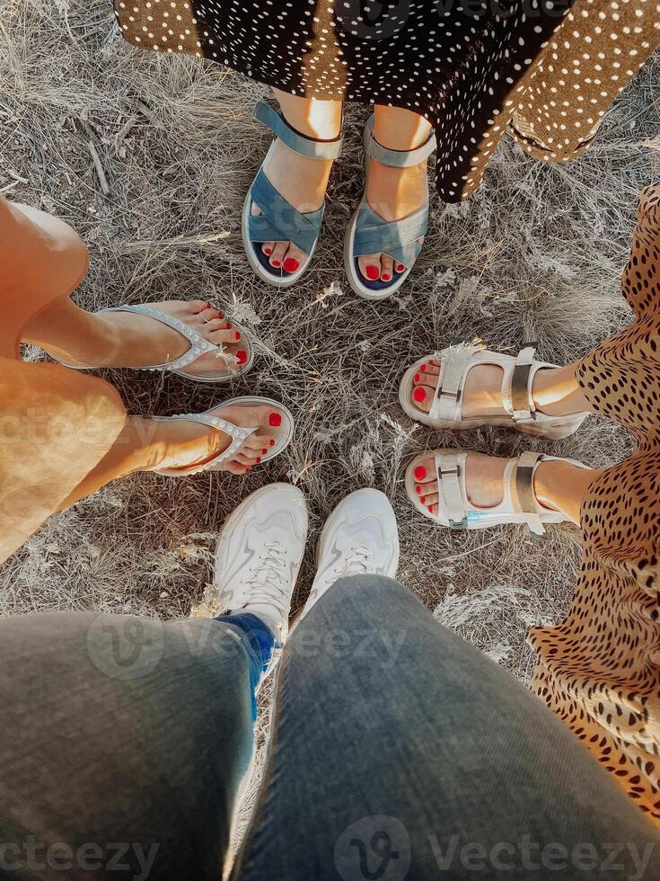 Four pairs of feet standing together on dry grass. Cute selfie of four couples female legs with red pedicure in white sandals and sneakers. Friendship and travel concept. photo