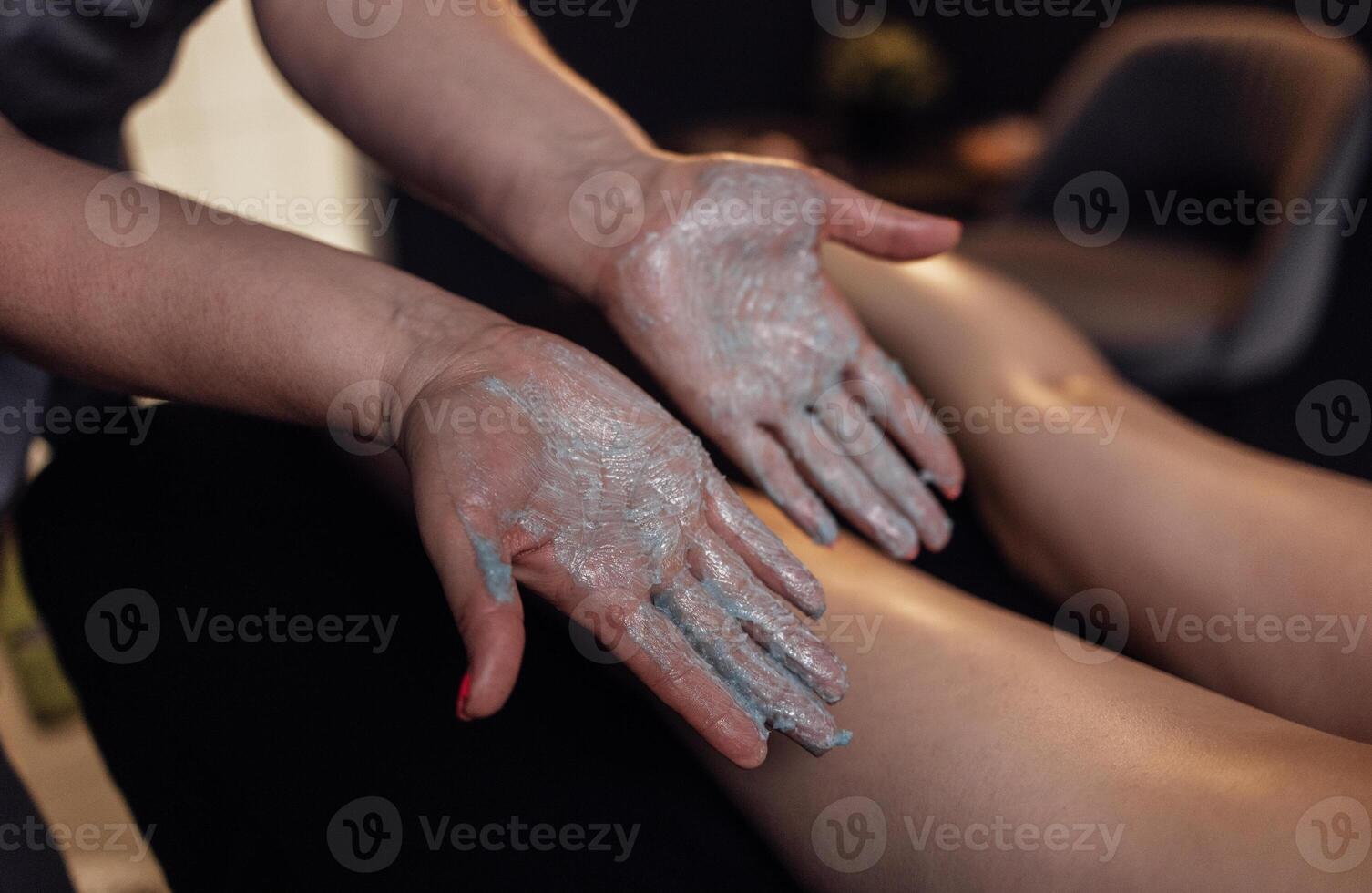 Close up of the masseuse palms with a grey scrub on them. Massage therapist applying cream to the female client legs. photo