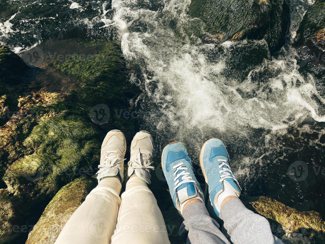 Feet in sneakers on the rocks, selfie over a fast river in the background. Top view of female and male legs over bubbling water. Tourists on a hike. photo