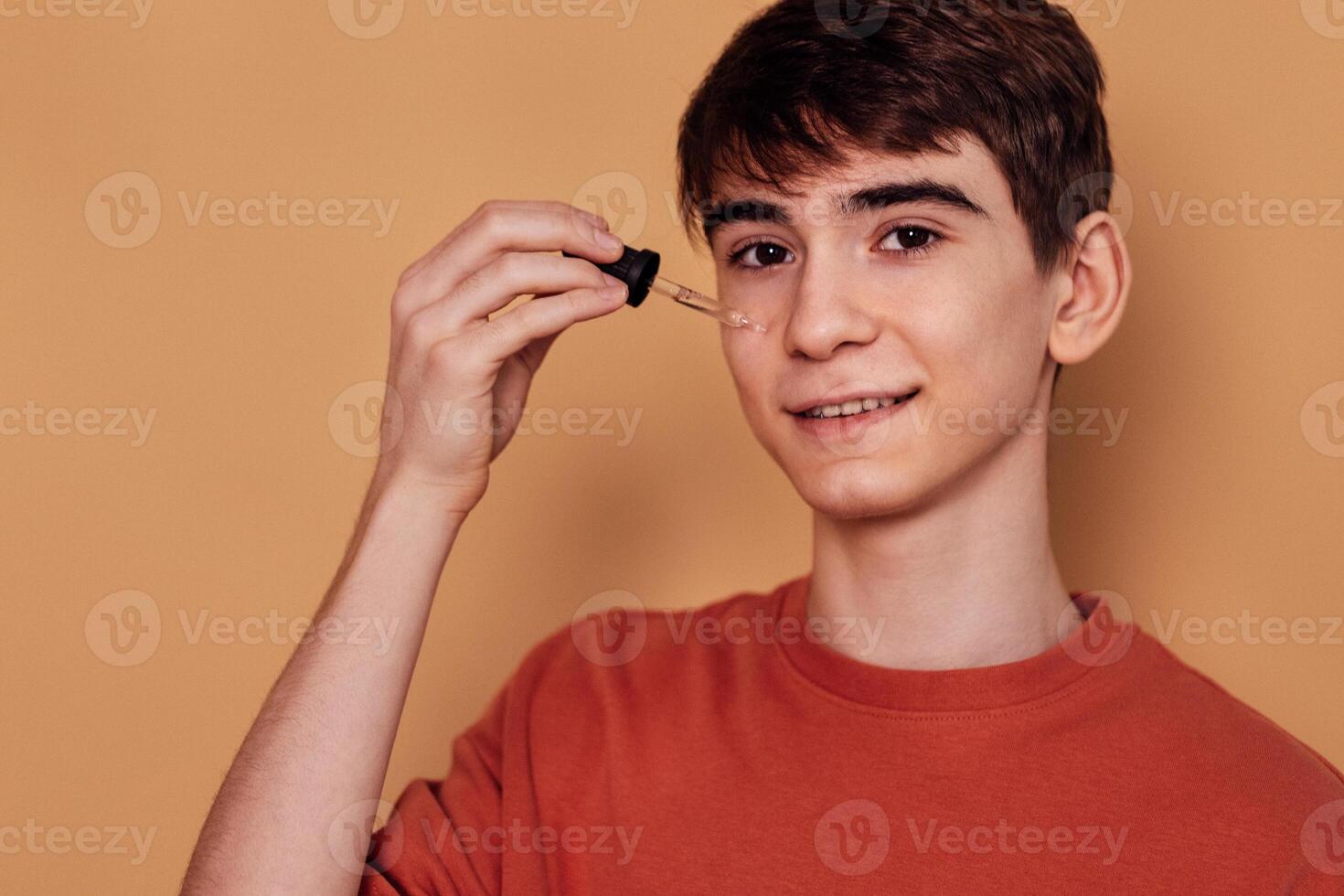 Teenage boy applying cleansing serum against acne and pimples on his cheek. Male teen in casual t-shirt is smiling and holding pipette. Hygiene and skin care in adolescence. photo