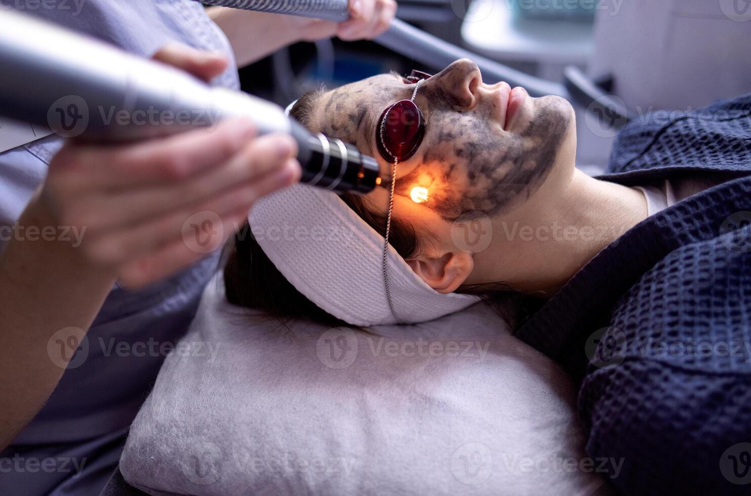 Beautician holding beauty laser near woman face covered with carbon mask photo