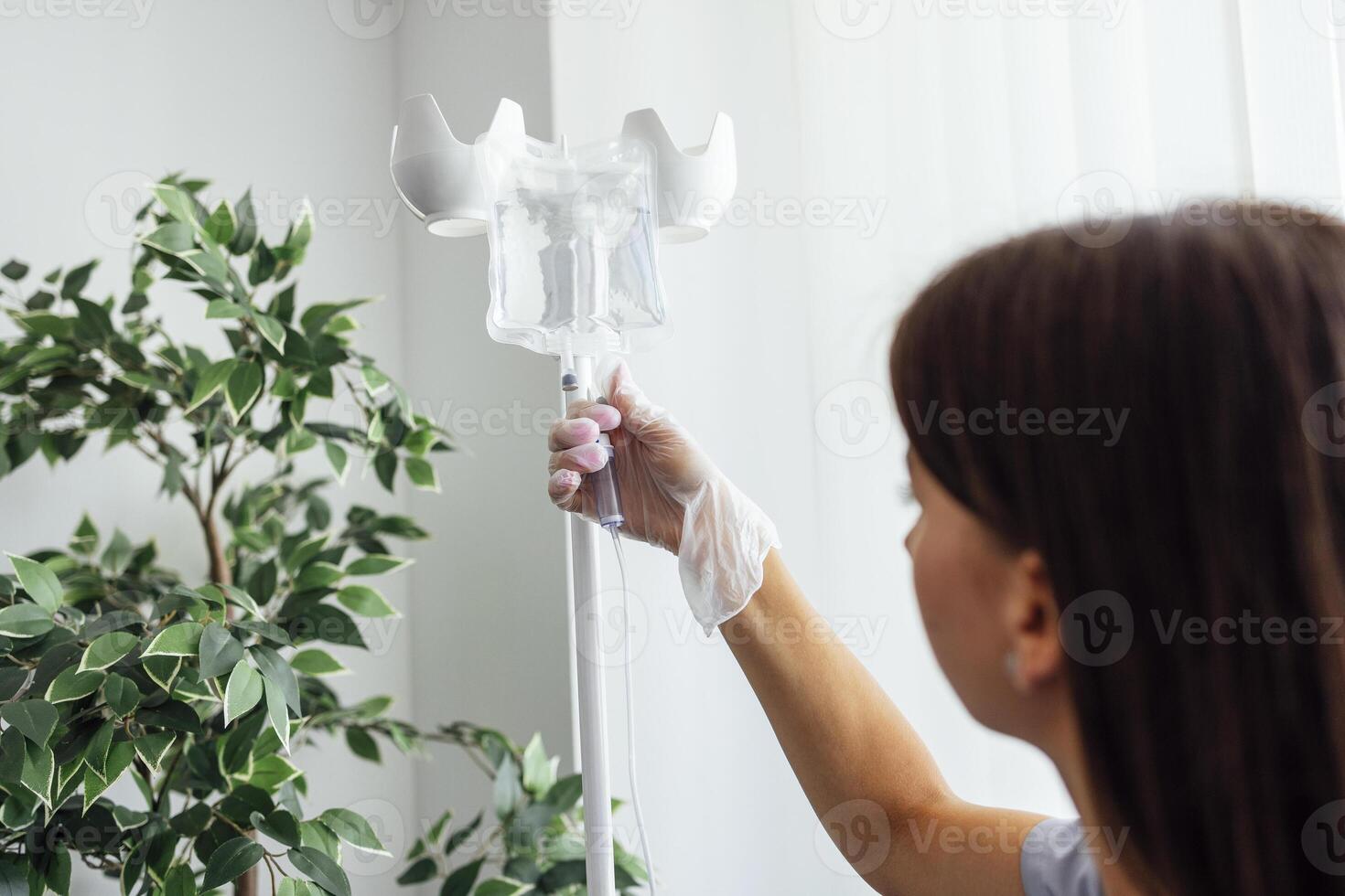 Young brunette nurse in transparent gloves installs a drip. Light room in the clinic. photo