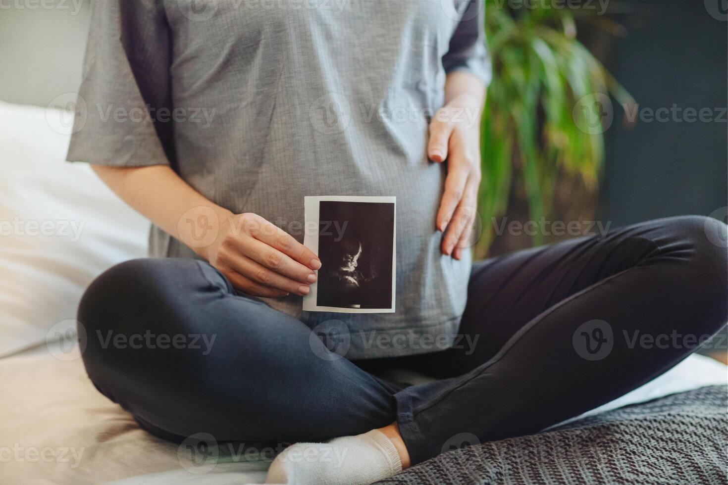 Cropped shot of pregnant woman holding pregnancy ultrasound while sitting in lotus pose on bed at home photo