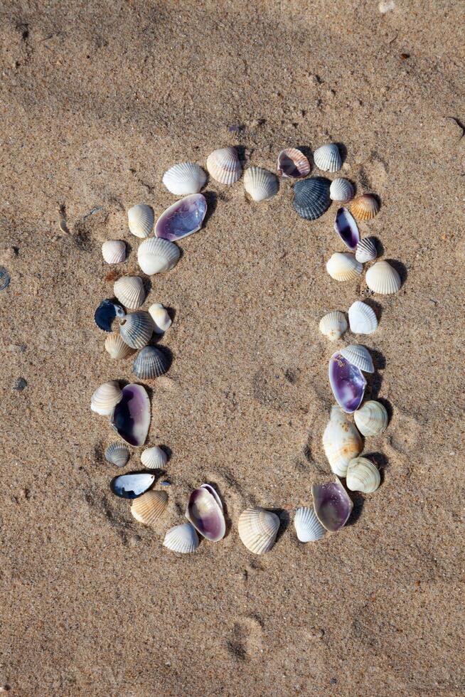 Number 0 laid out from seashells on the sand. Marine background. The symbol is zero. Shells. Copy space. photo