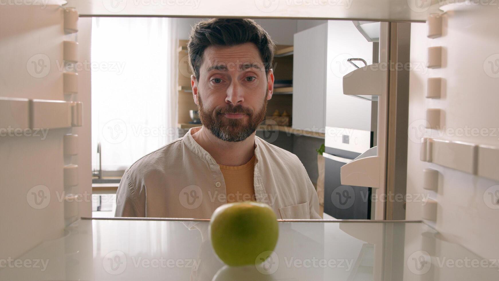 POV point of view from inside refrigerator Caucasian adult man at kitchen open empty fridge with one green apple fruit hungry guy male upset need food delivery meal crisis diet shrugs sad frustrated photo