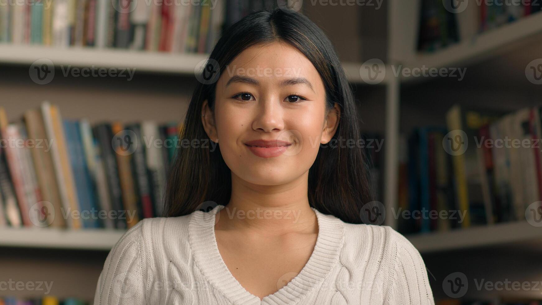 Young asian woman student posing in university library or bookstore with bookshelf in background ethnic smart girl bookworm reader smiling looking at camera female portrait college education concept photo