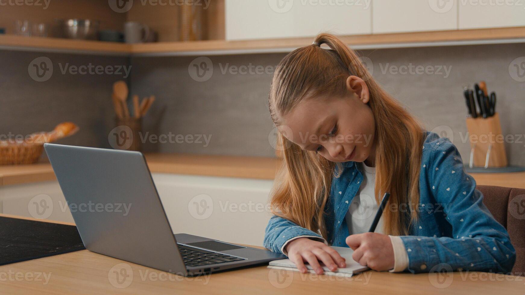 Caucasian kid girl school pupil studying online from home watching laptop web class lesson teacher by call e-learning pandemic isolation writing homework task at kitchen child remote education photo