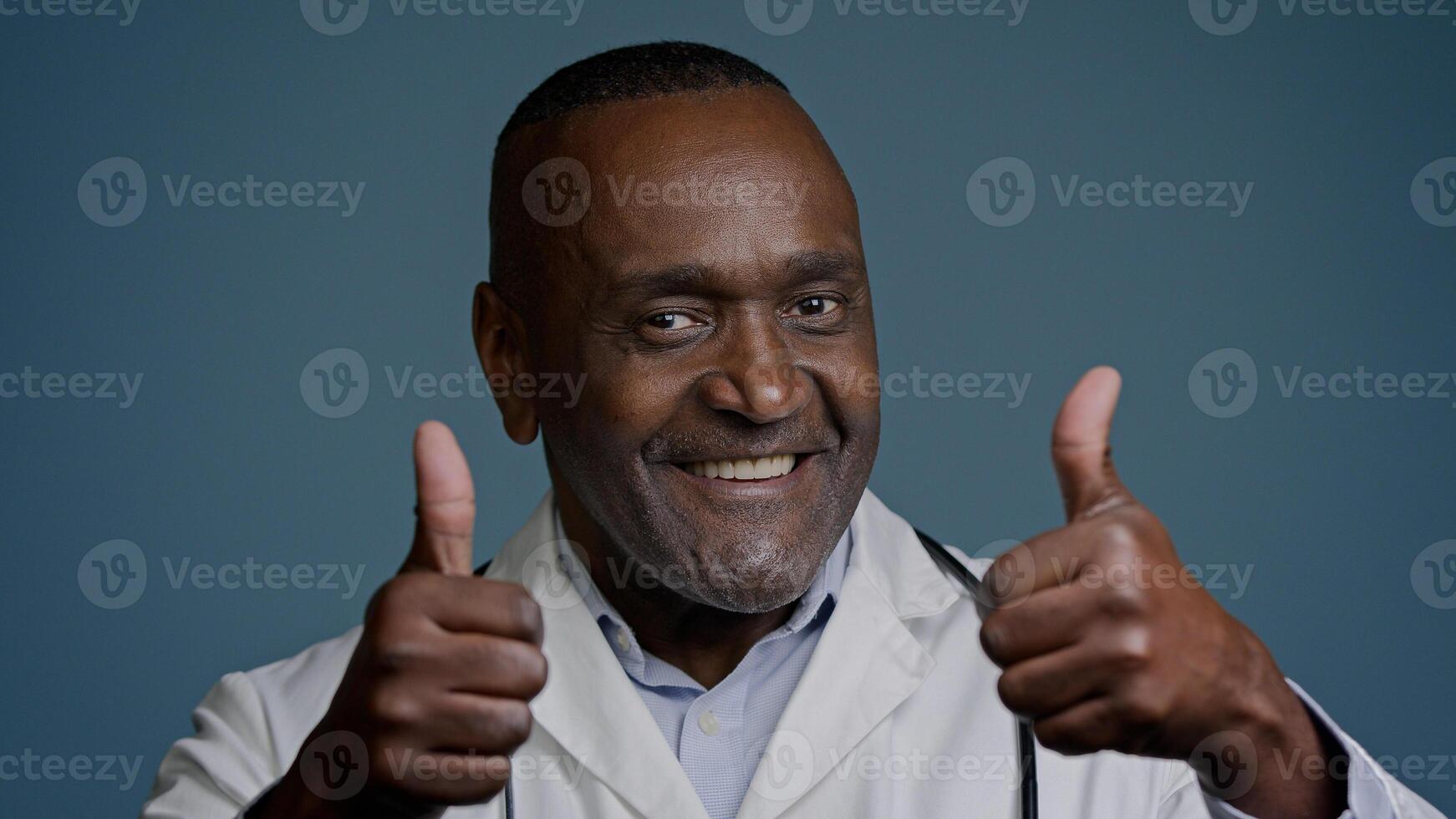 Portrait smiling African American man doctor in white coat showing thumbs up in studio blue background looking at camera gesture approval recommendation medical services health worker show like sign photo