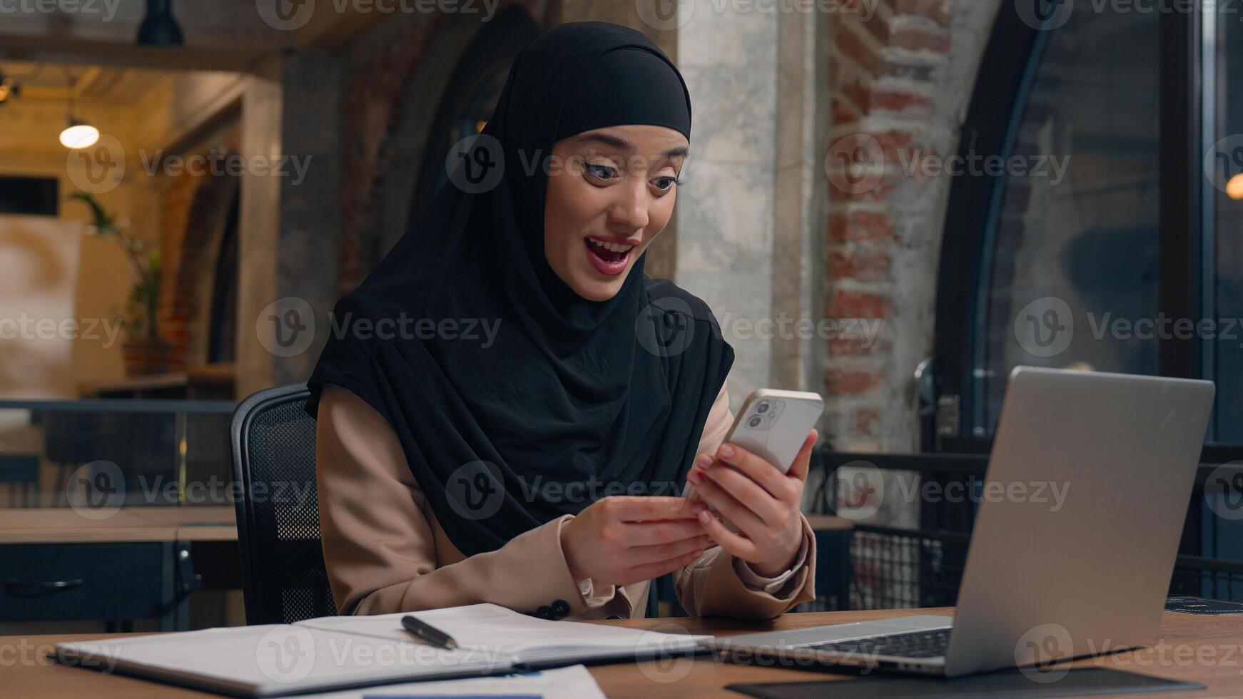 Happy excited joyful Arabian muslim business woman wonder amazement female islamic girl hijab user student businesswoman mobile phone good news achieve victory online bet shock win smartphone office photo