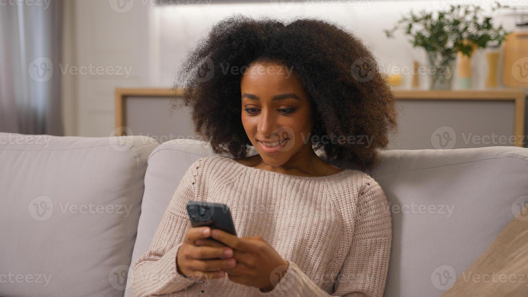 Happy African American girl smiling ethnic female woman using mobile phone cell gadget addicted chatting on smartphone shopping online on home sofa scrolling social media addiction influencer blogger photo