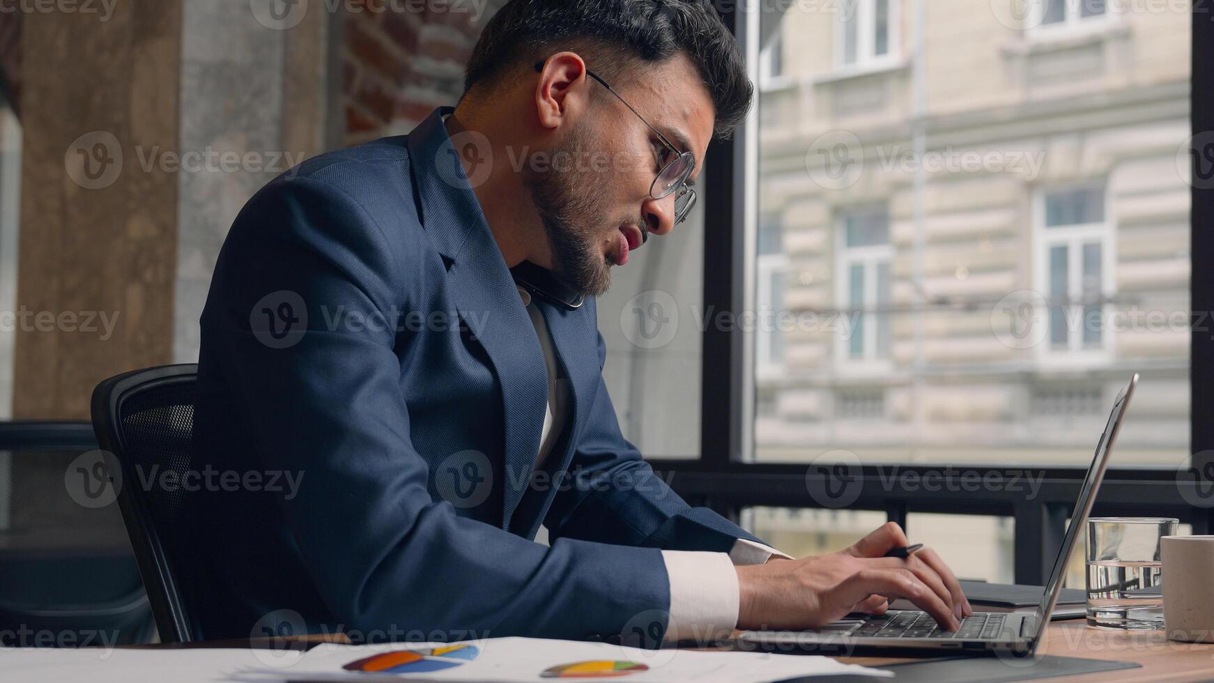 Stressed multitasking businessman Indian Arabian male guy annoyed employer manager consultant talking mobile phone shout working laptop writing documents anxious business man problem overwork office photo