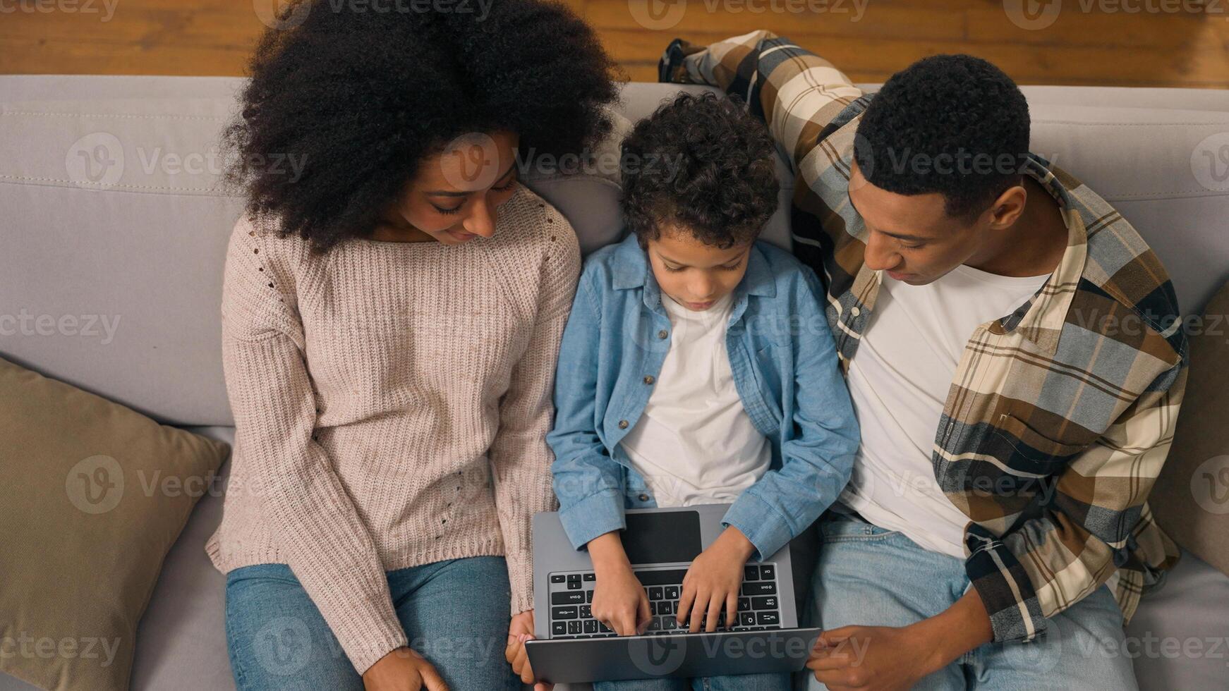 Top view African American family married couple teaching little boy child son kid offspring typing using tech laptop notebook pc device discuss speaking tell online education indoors living room couch photo