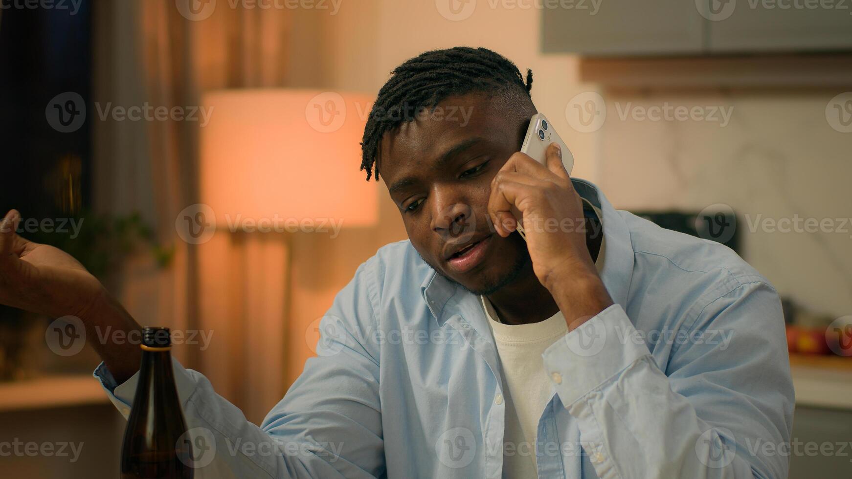 Drunk alcoholism addicted African American man guy male drinking alcohol drink beer at home evening talking mobile phone call smartphone talk speak suffering problem trouble sad upset stress depress photo