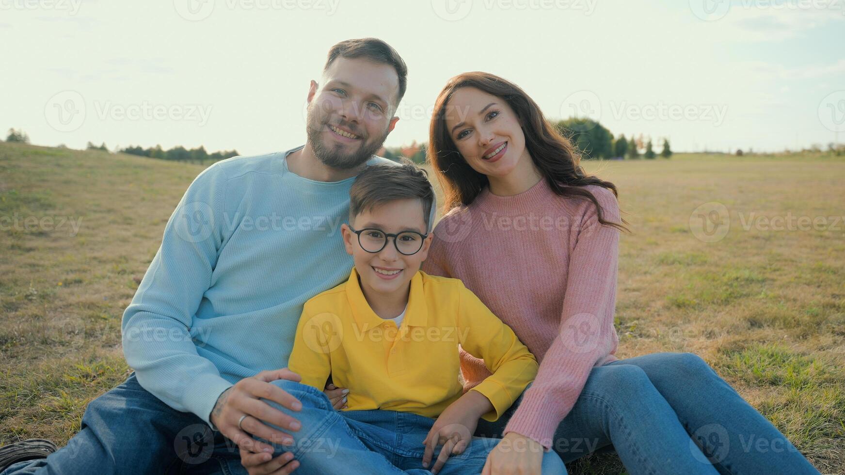 Cute Caucasian happy family portrait joyful man male dad smiling woman female mom little boy child kid son sitting together hug embrace looking camera outside nature field farmland park picnic nature photo