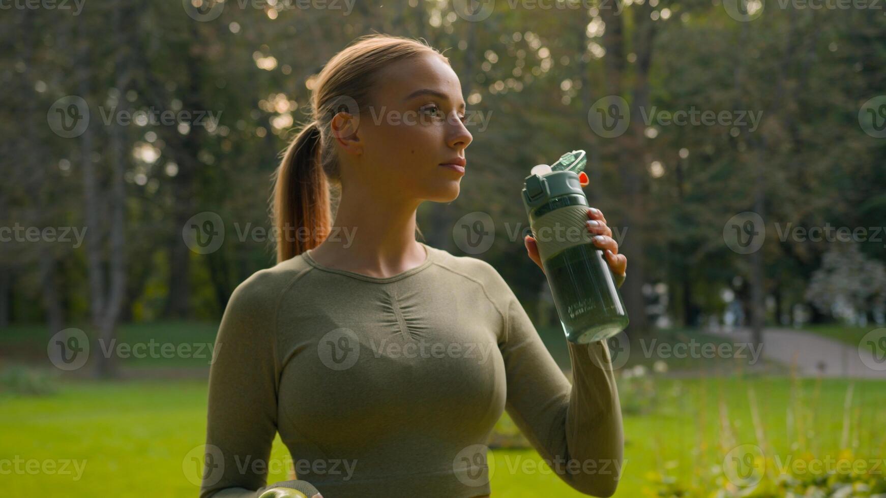 Sporty athletic thirsty girl Caucasian woman runner jogger drinking water after workout in park outdoors drink sport bottle healthy habit active young lifestyle refreshment hydration smiling to camera photo